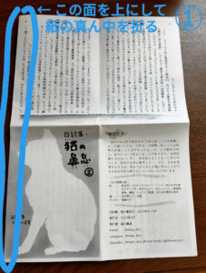 ネットプリントに登録した日記ZINEを印刷したあとの折り方・綴じ方・裁ち方の手順（1）です。 まず、猫の絵の面が上にくるようにして、長辺を半分に折ります。