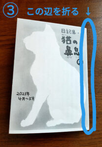 ネットプリントに登録した日記ZINEを印刷したあとの折り方・綴じ方・裁ち方の手順（3）です。 次に、猫の絵の面と、あとがきの面の真ん中を折ります。